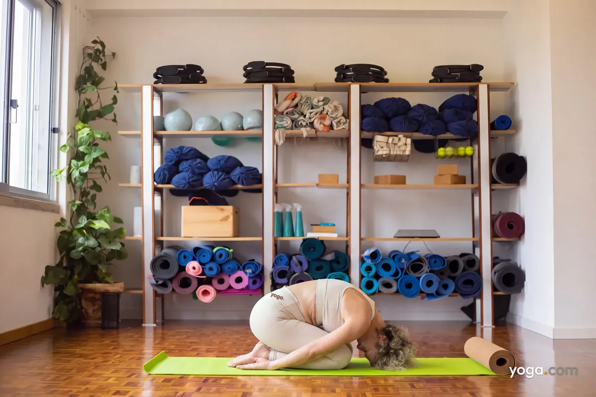 A woman wearing a beige matching set rests in child’s pose on a green mat inside a yoga studio.