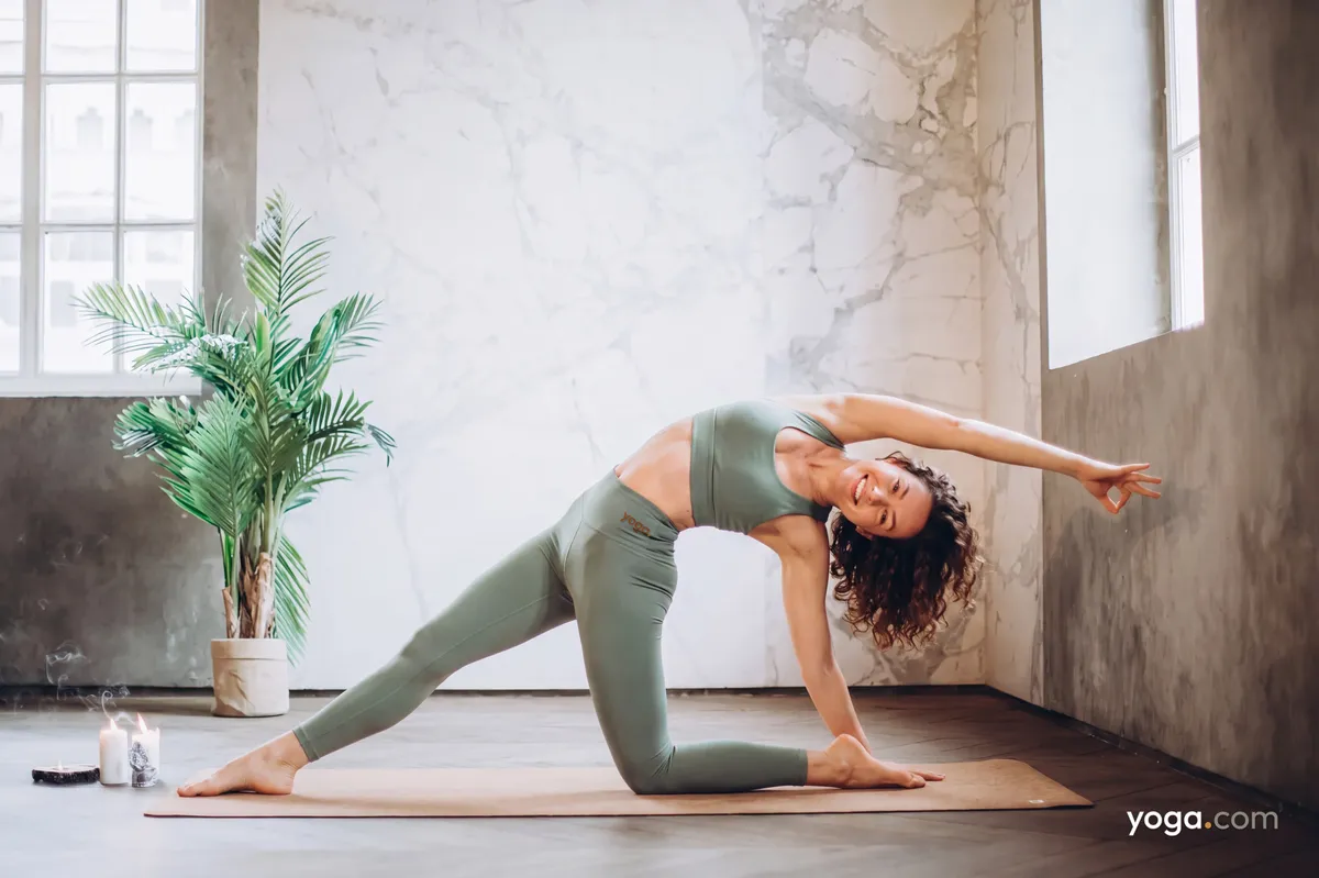 A woman in sage green activewear practices a backbend yoga pose on a mat in a minimalist home corner.