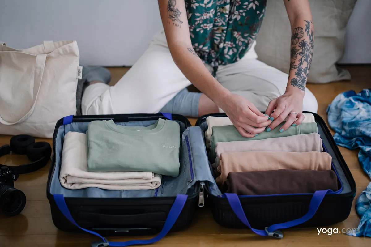 A person with tattooed arms packs folded and rolled neutral-toned clothes into an open suitcase on a wooden floor.