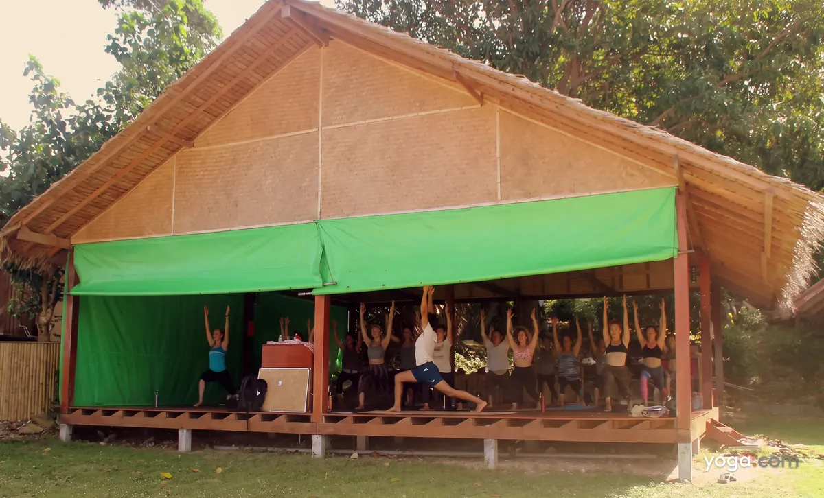 People in activewear practice a warrior yoga pose inside an open-air wooden pavilion with a thatched roof.