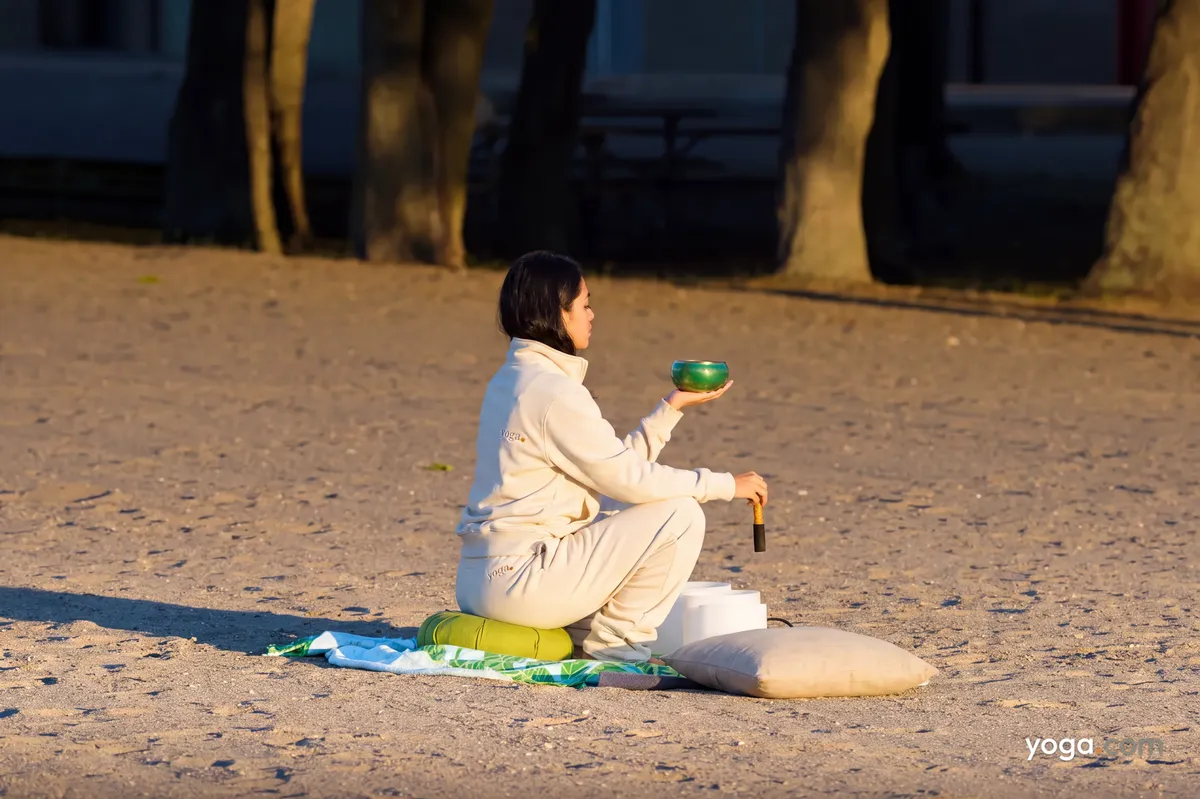 A woman in a cream tracksuit squats on a sandy beach, holding a green singing bowl for outdoor meditation.